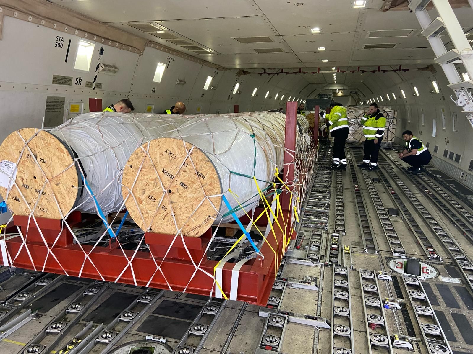 Loadmasters in high-visibility jackets checking the netting over two large cable reels secured inside a cargo aircraft.