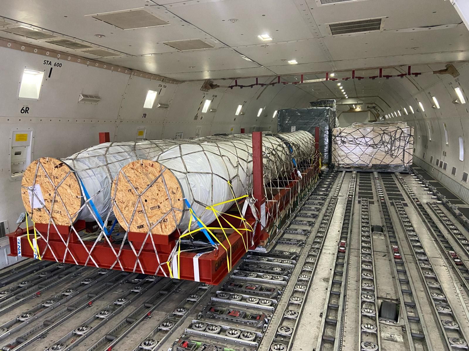 Two large timber cable reels and a wrapped tubing bundle netted onto pallets inside the main deck of a freighter aircraft.