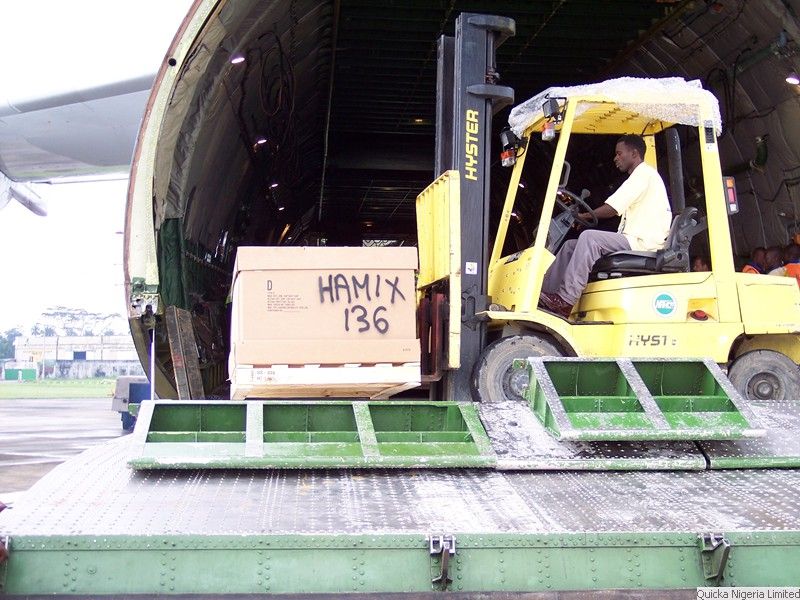 A forklift carrying a large timber case marked HAMIX 136 up a ramp into the open nose door of a cargo aircraft.
