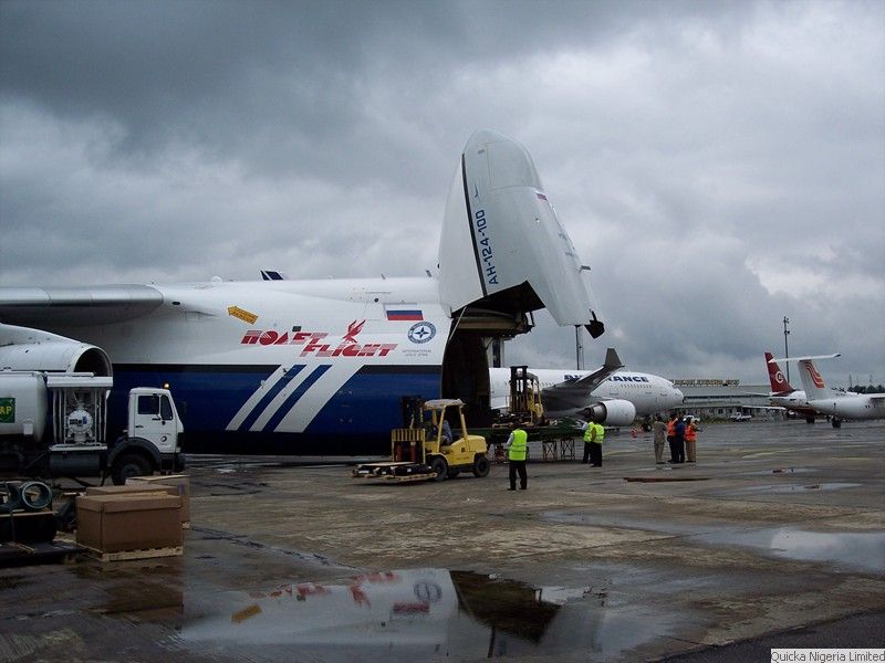 A heavy-lift cargo aircraft with its nose door open on the apron, a forklift and truck working alongside it, and other aircraft parked beyond.