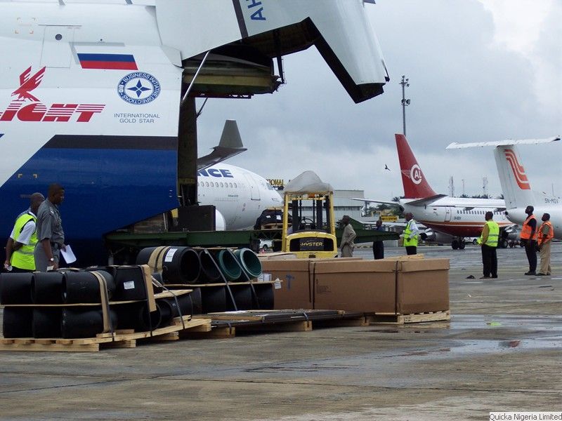 A heavy-lift freighter aircraft with its nose door raised, cargo and timber cases on the apron in front of it, and handlers in orange high-visibility vests working alongside.