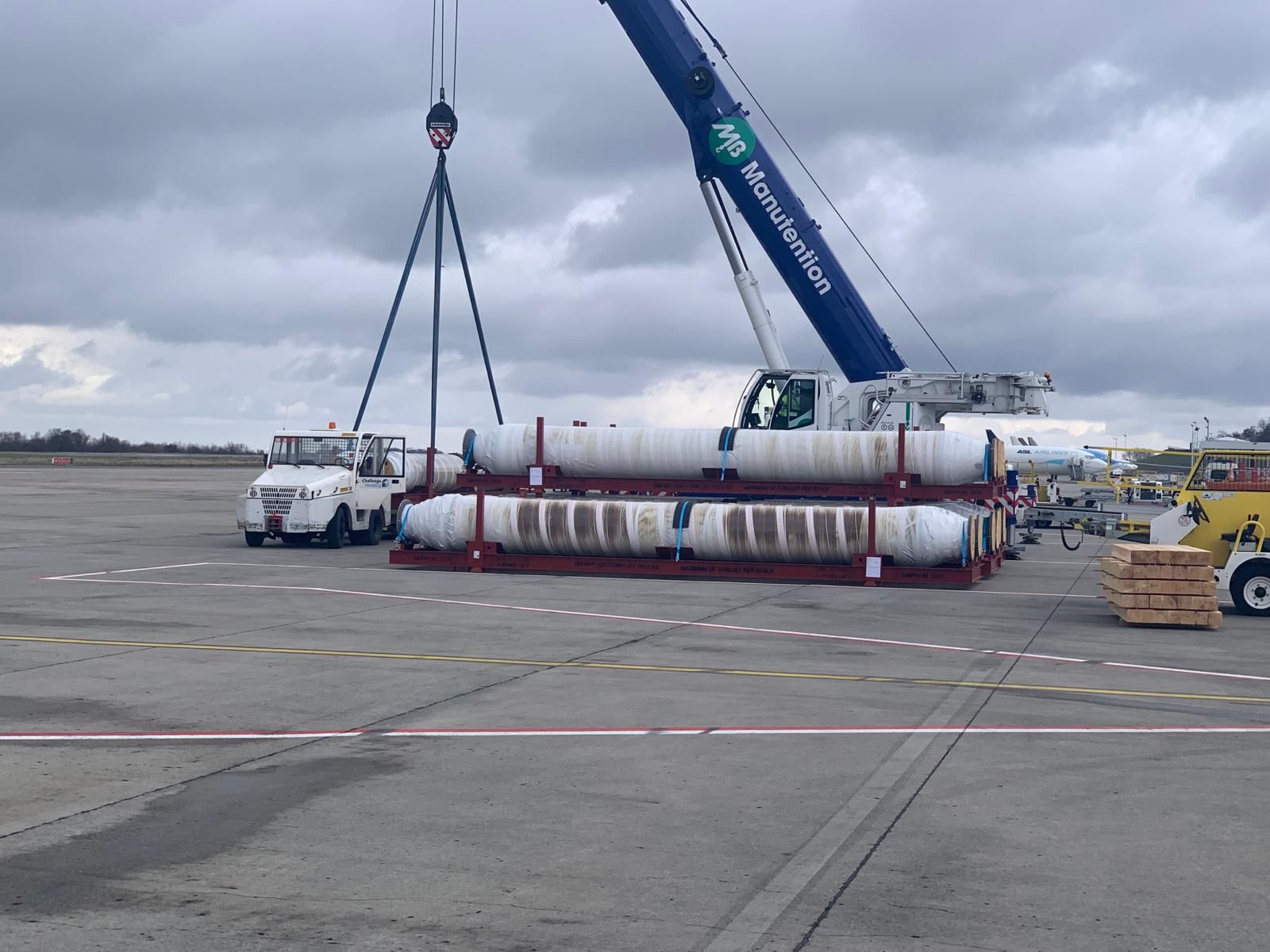 A mobile crane lifting long wrapped tubing bundles on a red pallet beside a cargo aircraft, on a wet apron under grey cloud.