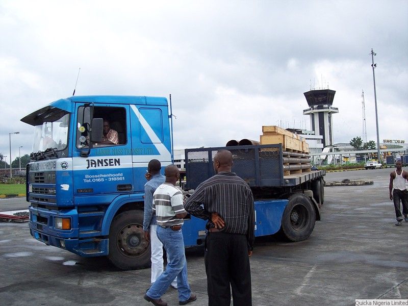 A blue flatbed truck loaded with palletised cargo on the airport apron, with three men in discussion in the foreground and the control tower behind.