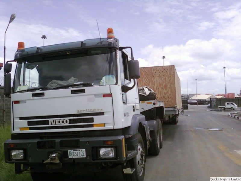 A truck carrying a large timber case on the apron of an airport, photographed from the front.