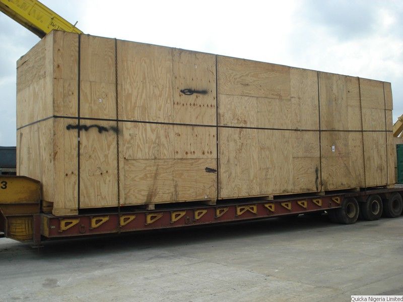A very large plywood case strapped to a trailer at a port, with a crane hook and slings above it.