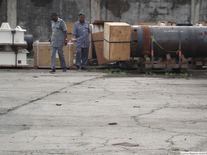 Two officials in uniform standing beside timber cases and a large steel vessel on a concrete apron during a cargo examination.