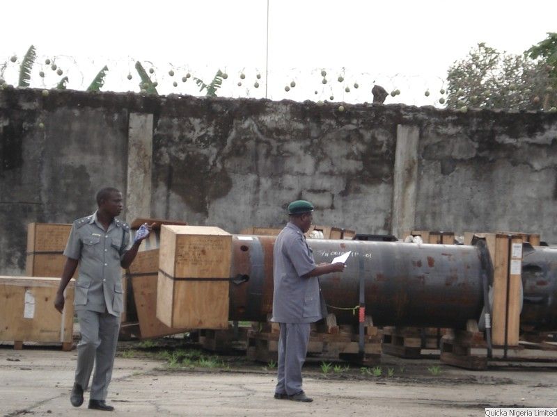 Two uniformed officials reviewing documents beside timber cases and a steel vessel during a cargo inspection.