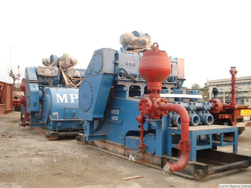A large blue and red drilling mud pump unit standing on a quayside, with pipework and a red structural frame beside it.