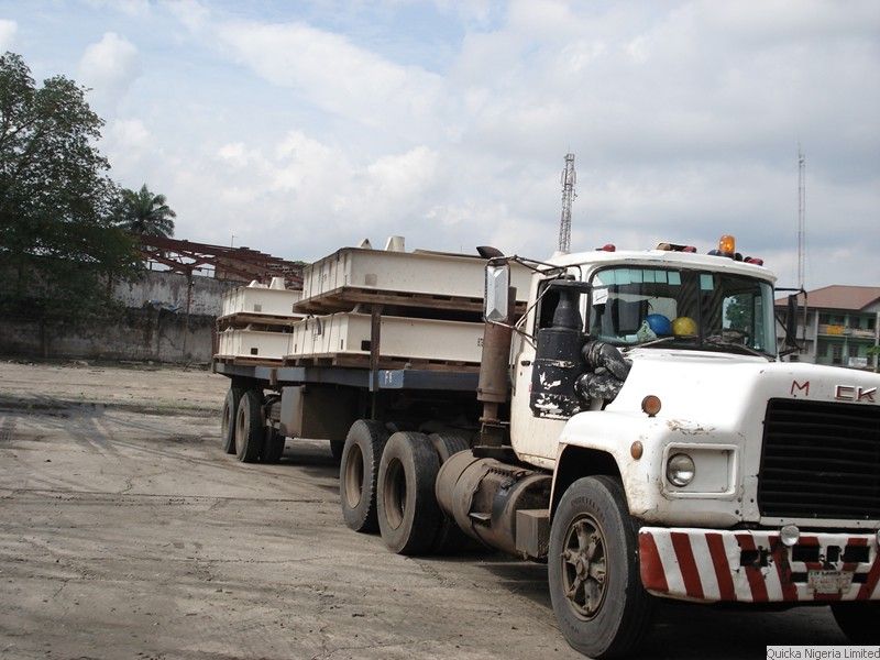 A Mack tractor unit coupled to a flatbed trailer loaded with structural steel sections, in a port yard.