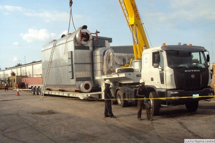 A large industrial boiler loaded on a low trailer behind a cream tractor unit, with a mobile crane alongside and crew supervising.