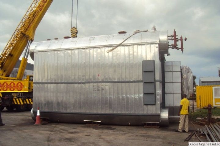 A large clad industrial boiler standing on timber packers in a yard, with a mobile crane rigged alongside it.