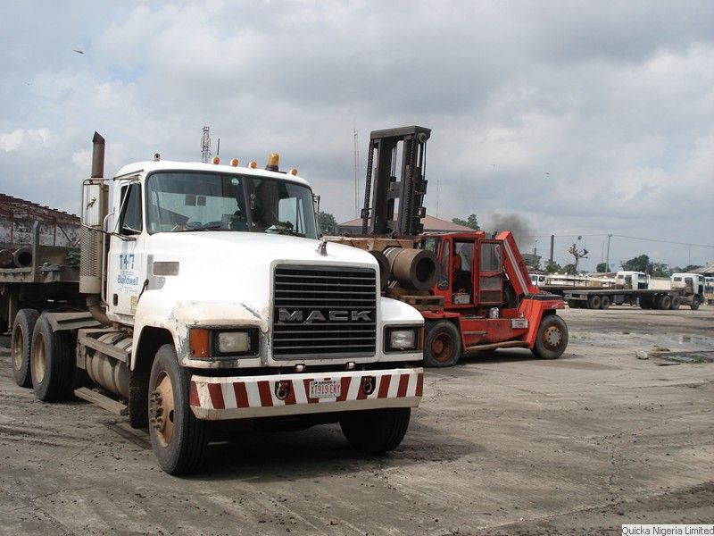 A white Mack tractor unit parked in a port yard with forklifts and other trucks behind it.