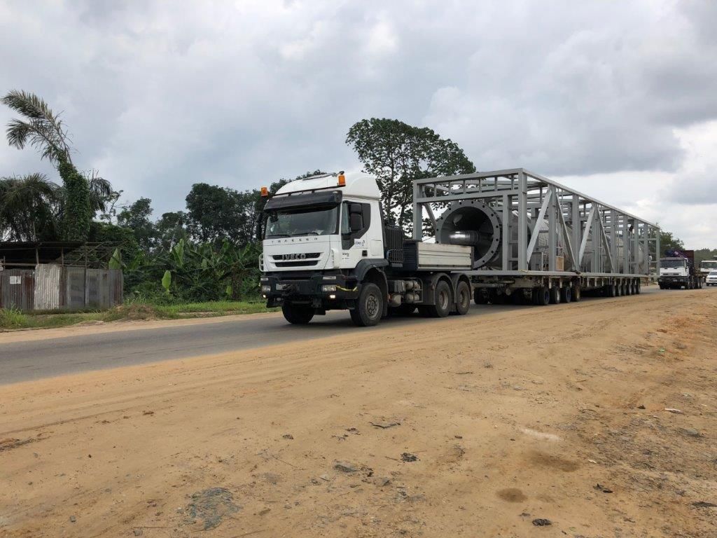 A tractor unit hauling a pressure vessel in an open steel frame on multi-axle trailers along a tree-lined highway, seen from the roadside.