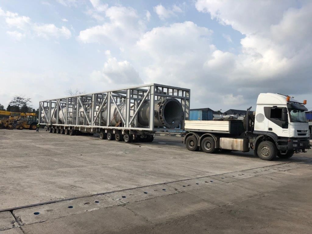 Large cylindrical pressure vessels carried in an open steel transport frame on multi-axle trailers, coupled to a white tractor unit on a concrete hardstanding, with wheeled plant and containers behind.
