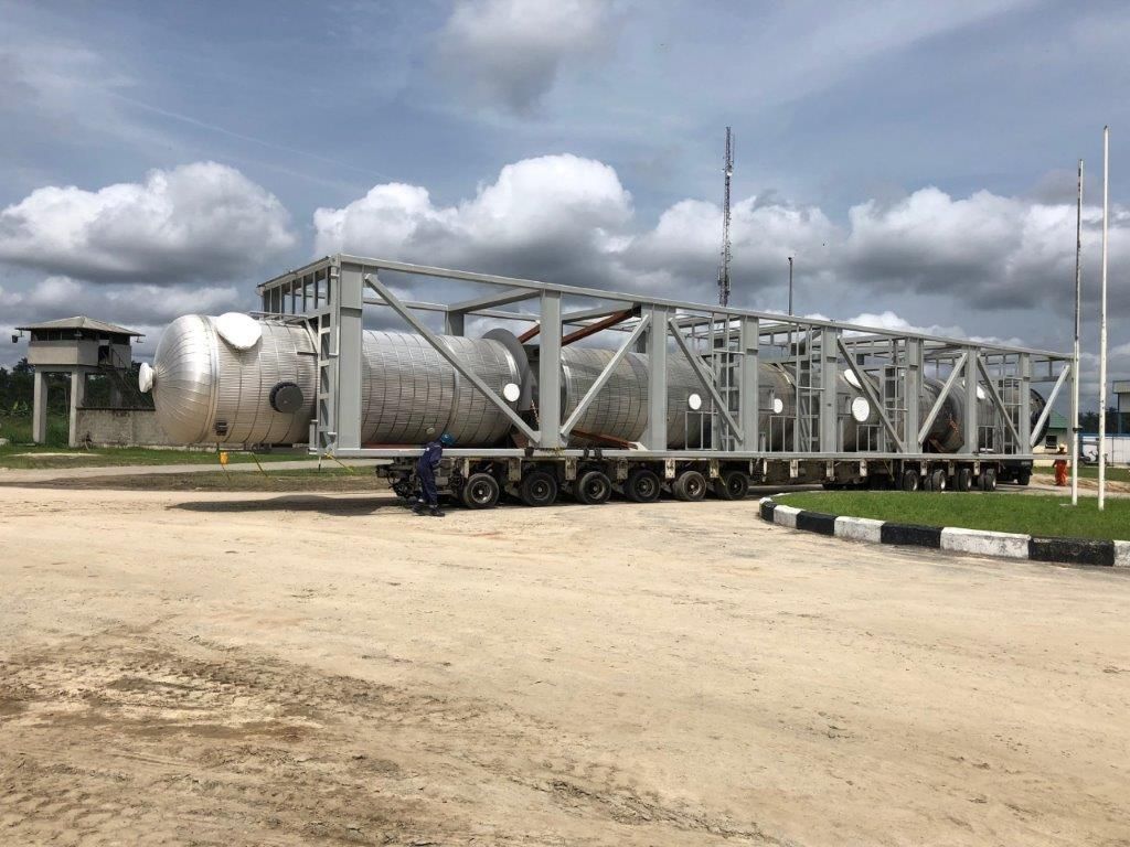 Several large clad cylindrical pressure vessels in a steel transport frame on multi-axle trailers, arriving on a prepared site with a gatehouse, a lattice mast and flagpoles behind.
