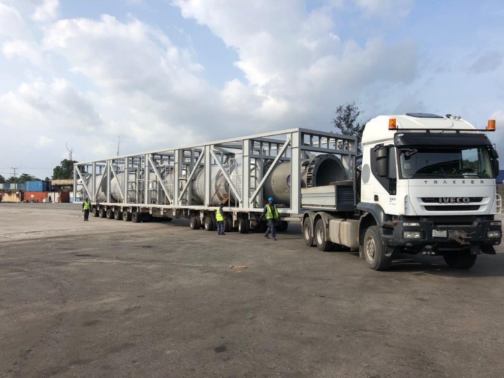 Cylindrical pressure vessels in an open steel transport frame on multi-axle trailers, with two men in high-visibility vests and hard hats standing alongside.