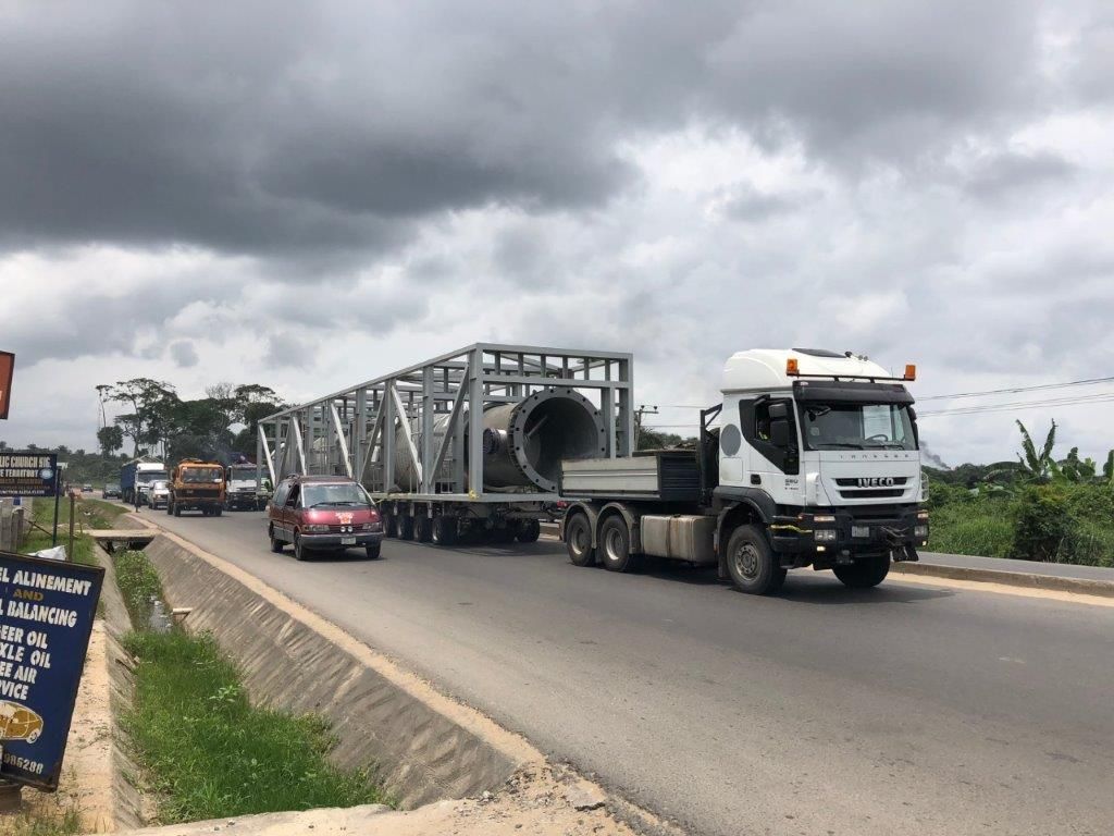 A cylindrical pressure vessel in an open steel space-frame on multi-axle trailers, travelling along a public road in Nigeria with cars and a lorry in the opposite lane.
