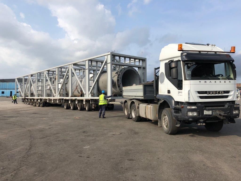Cylindrical pressure vessels in an open steel transport frame on multi-axle trailers in an open yard, with a banksman in a high-visibility vest signalling beside the load.