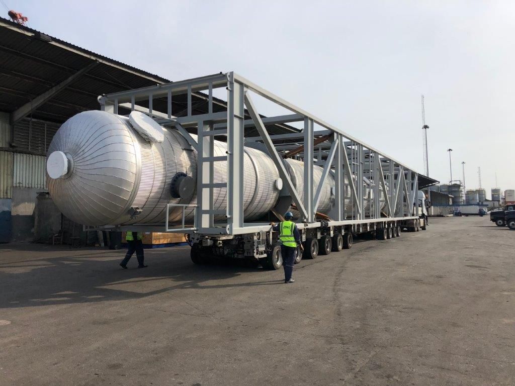 Two large polished pressure vessels mounted in a steel transport frame on multi-axle trailers, under a warehouse canopy.