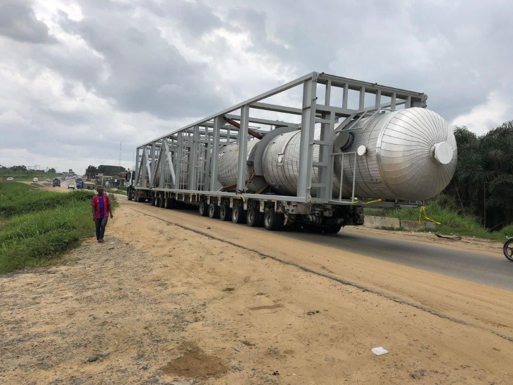 A long multi-axle trailer carrying framed pressure vessels along a road, with a man walking on the verge beside it.