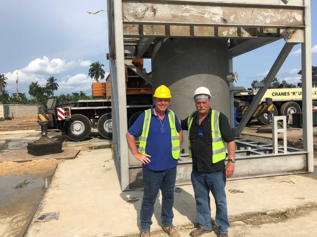Two men in hard hats and high-visibility vests standing at the base of the installed tower, with a crane behind them.