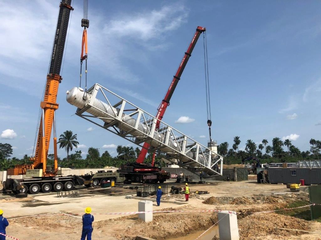 Two mobile cranes, one orange and one red, beginning to raise a tall steel process tower from horizontal on a prepared foundation.