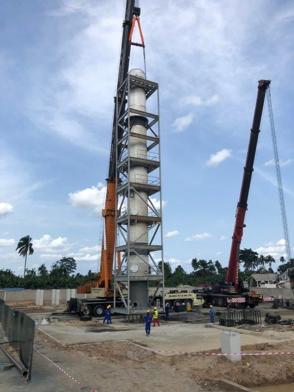 The vertical process tower held by two cranes directly over its foundation, with crew positioned at the base.