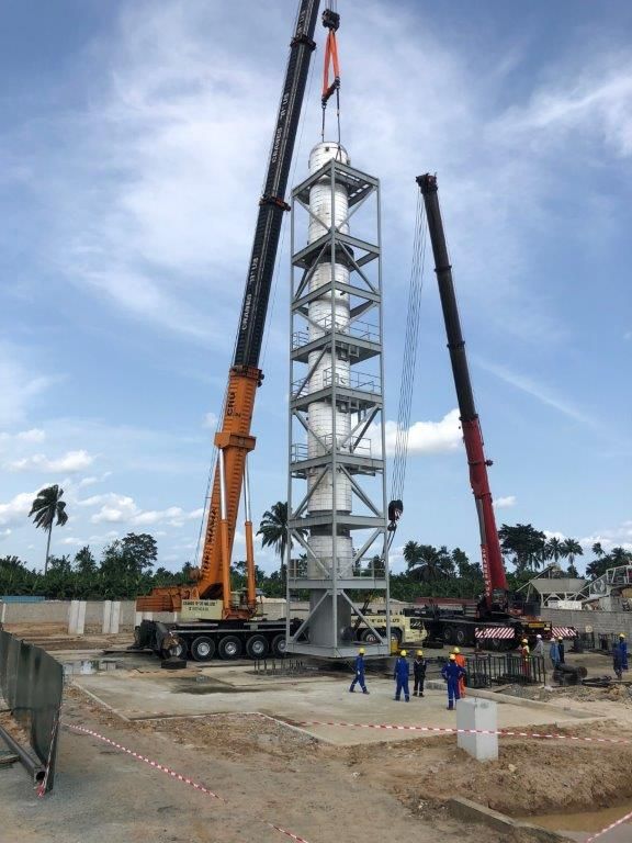 The process tower standing vertical between two mobile cranes, still slung, above its concrete foundation.