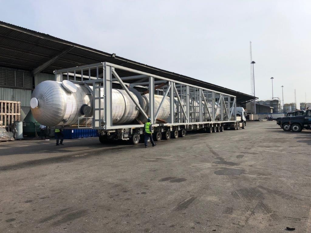 Pressure vessels in a steel frame on a long multi-axle trailer, parked under a warehouse canopy with a second truck beyond.
