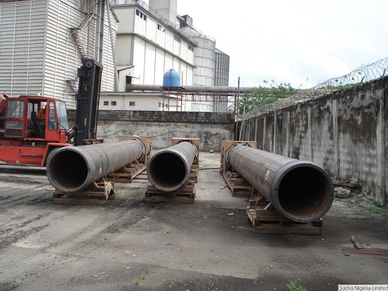 Three sections of large-diameter steel pipe resting on timber dunnage in a yard, with a forklift and silo structures behind.