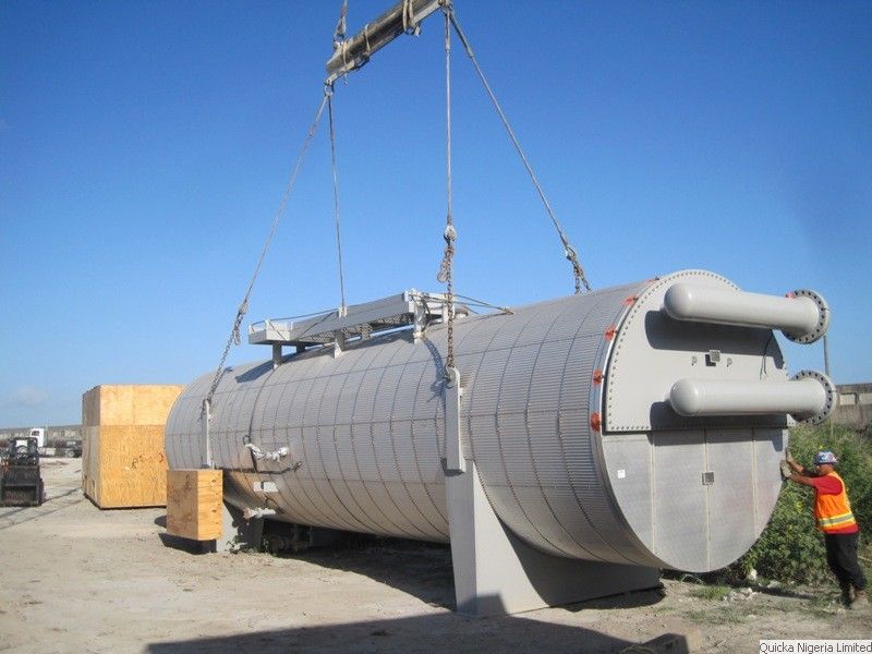 A large cylindrical vessel with two nozzles being lifted on slings by crane, with a banksman guiding it from below.