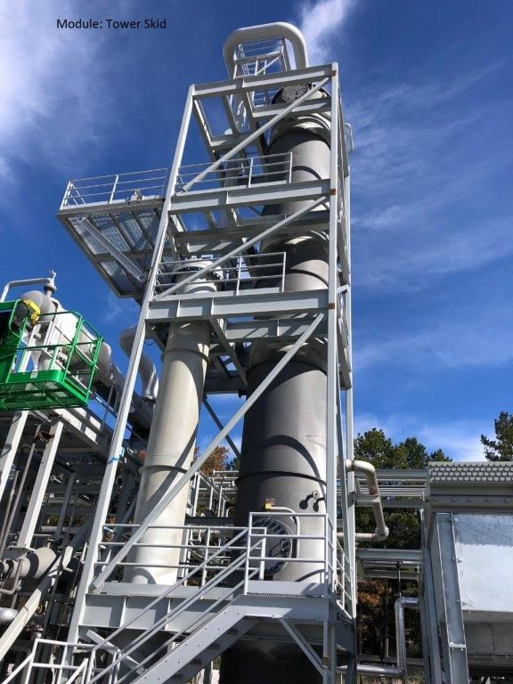 A tall tower skid with a vertical process column and access stairways, standing in a fabrication yard.