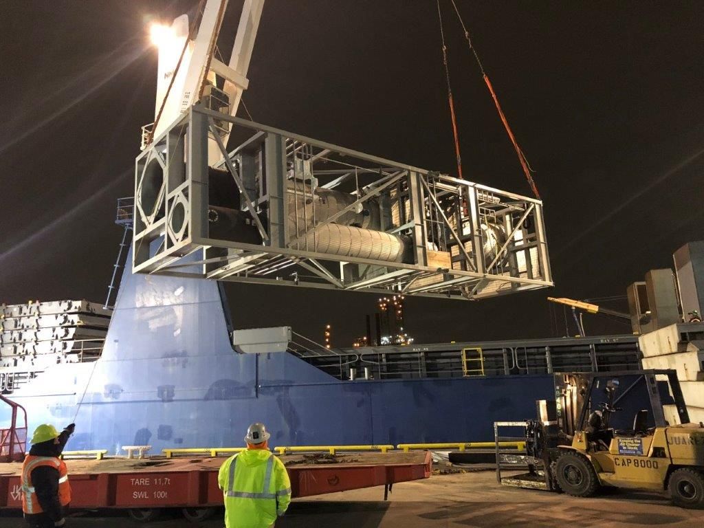 A steel drilling module being lifted by crane onto a barge at night under floodlights, with crew in high-visibility clothing on the deck below.