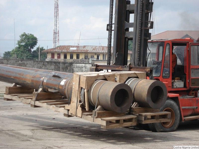 A heavy forklift handling large-diameter pipe sections resting on timber cradles in a port yard.