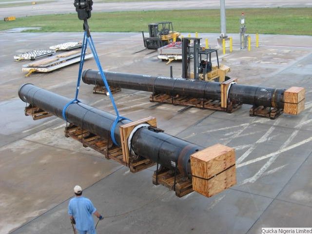 Two long steel pipe sections slung on blue strops being lifted above timber cradles on an airport apron, with aircraft pallet dollies and a ground-support forklift behind and a banksman below.