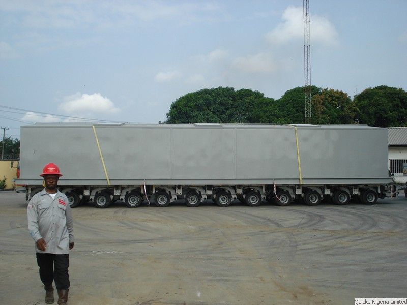 A very large grey power plant module carried on a self-propelled modular transporter, with a supervisor in a red hard hat walking alongside.