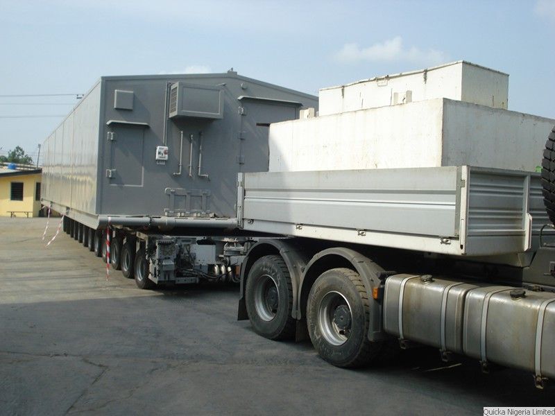 A large grey module on a modular transporter coupled to a heavy tractor unit, in a port compound.