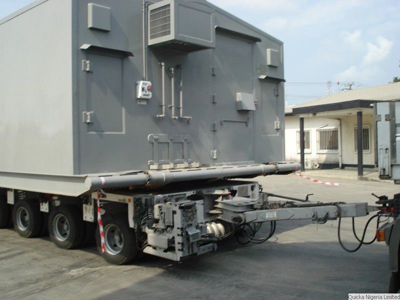 The drawbar and steering gear of a modular transporter carrying a large grey module, in a port compound.