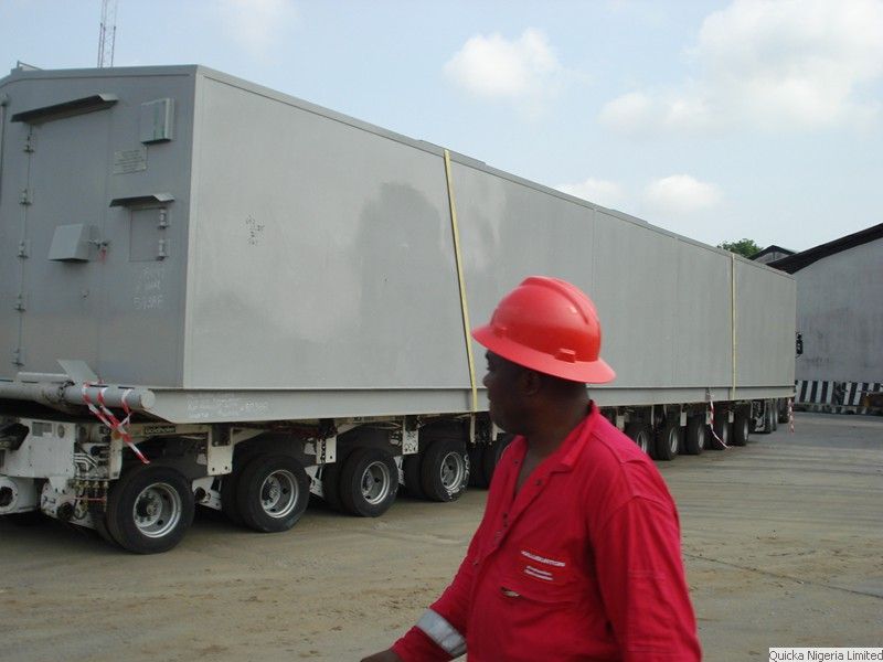 A supervisor in red coveralls and hard hat walking beside a large grey module on a multi-axle transporter.
