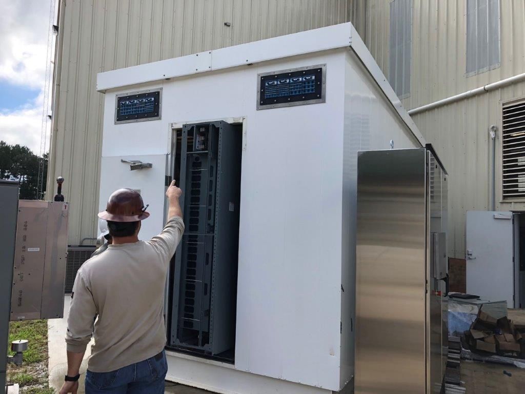 A man in a hard hat standing outside a small white prefabricated module at a fabrication yard, pointing into its open doorway at a rack of electrical equipment inside.