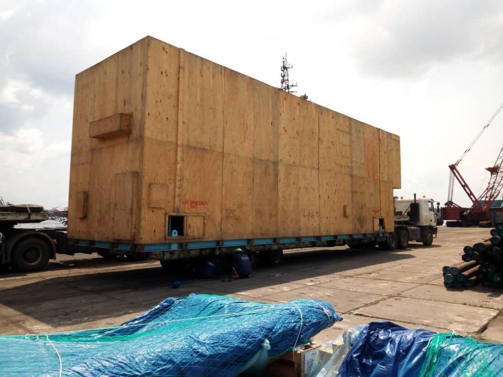 A very large timber case loaded on a multi-axle lowbed trailer at a port, with a crane and tarpaulins in the foreground.