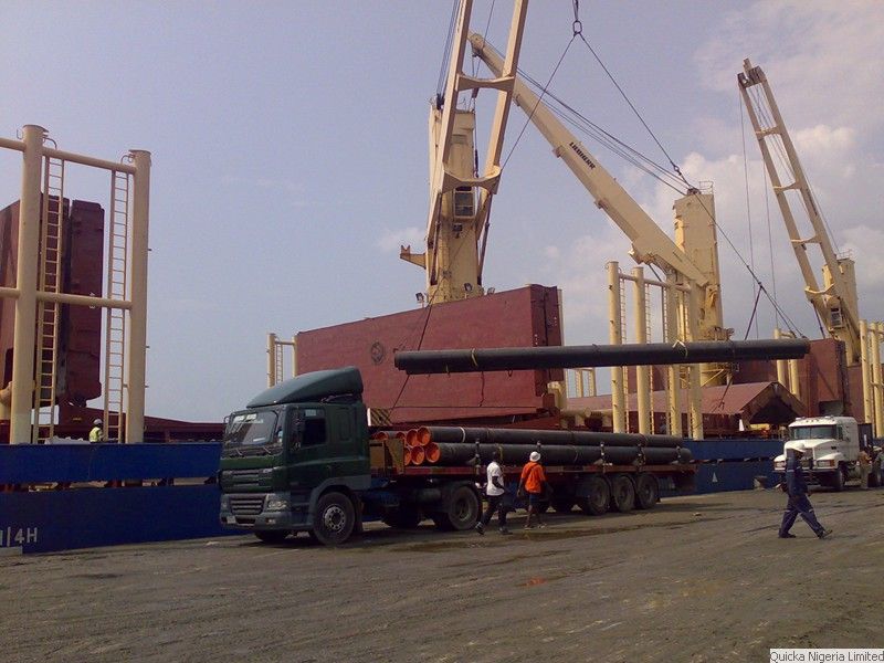 Long steel pipes being lifted by ship's crane onto a flatbed truck at a quayside, with stevedores working below.
