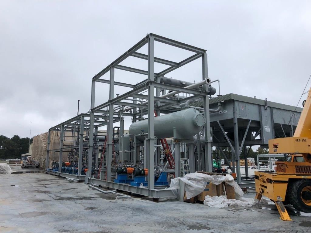 A row of steel-framed process modules with pipework and vessels, lined up in a fabrication yard with a crane beyond.