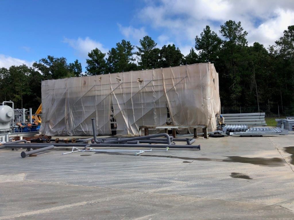 A large module fully shrink-wrapped in protective sheeting, standing in a fabrication yard with loose pipework on the ground nearby.