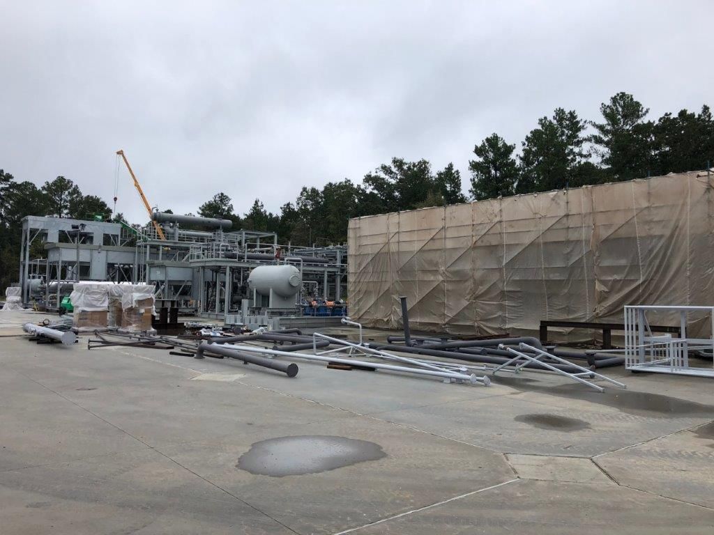 A wide view of a fabrication yard with several process modules, a wrapped module and stacked pipework under a cloudy sky.