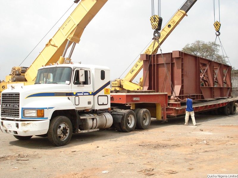 A white Mack tractor unit with a flatbed trailer carrying a large red structural steel frame, being loaded by crane at a port.