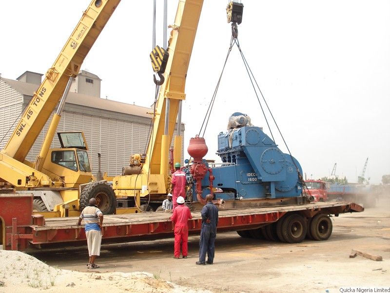 Two yellow mobile cranes lifting a large blue drilling unit onto a flatbed trailer at a port, with crew in red coveralls standing by.