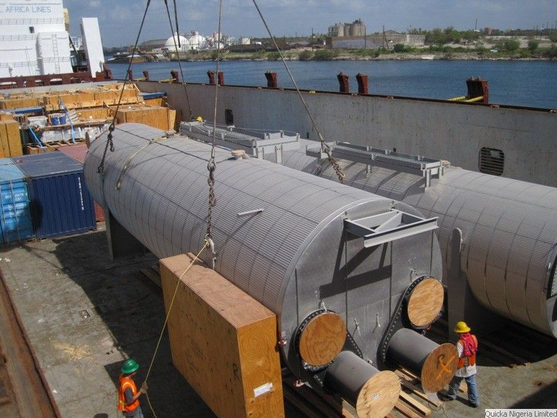 Two large clad cylindrical vessels resting on timber dunnage beside a timber case being lifted on wire ropes, with a ship’s side lettered AFRICA LINES and a waterfront behind, and crew in hard hats and high-visibility vests alongside.
