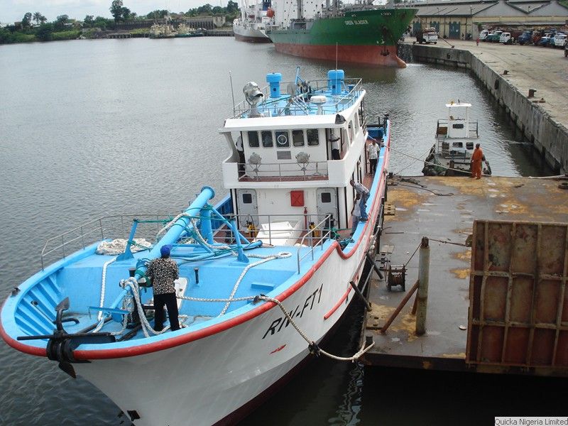 A blue and white workboat moored alongside a quay at Port Harcourt, with a cargo vessel and barge in the background.